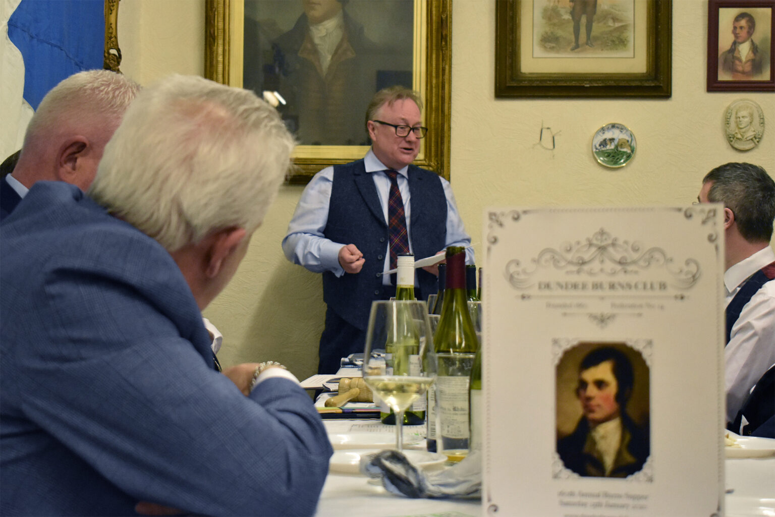A speaker stands at the head of the table beside a portrait of Burns