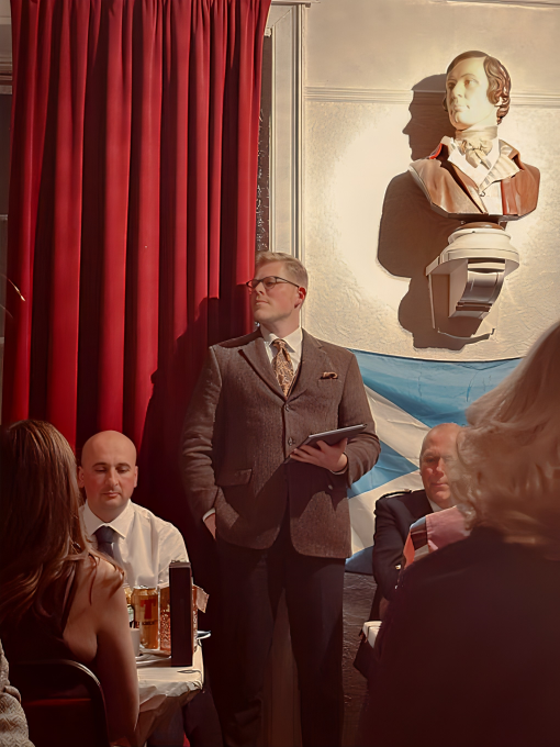 A speaker in a tweed suit stands beneath the bust of Burns