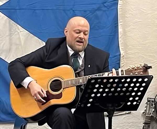 A musician sings with a twelve-string guitar in front of a Saltire
