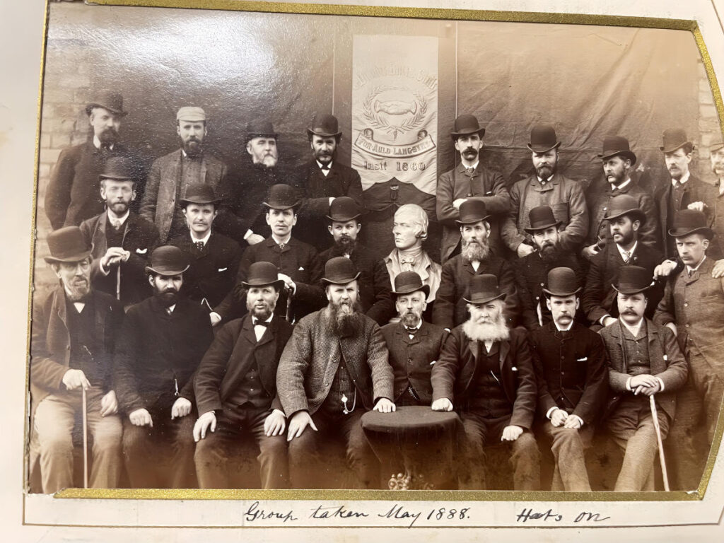 Around thirty Dundee Burns Club members in hats and overcoats posed around a bust of Burns, with the club banner behind